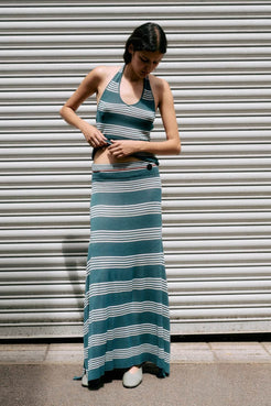 A woman wearing a teal and white striped halter-neck maxi dress with a matching cropped top, standing in front of a gray metal shutter.