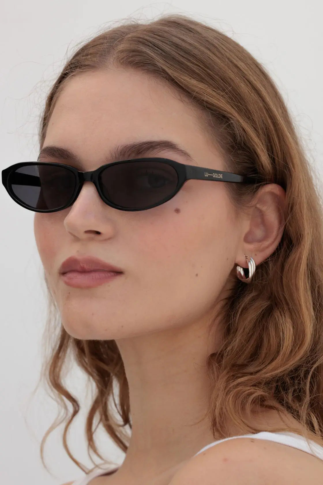Model wears fitted black oval sunglasses with a contoured silhouette, styled with a natural beauty look for versatile warm-weather outfits.