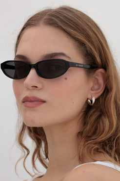 Model wears fitted black oval sunglasses with a contoured silhouette, styled with a natural beauty look for versatile warm-weather outfits.