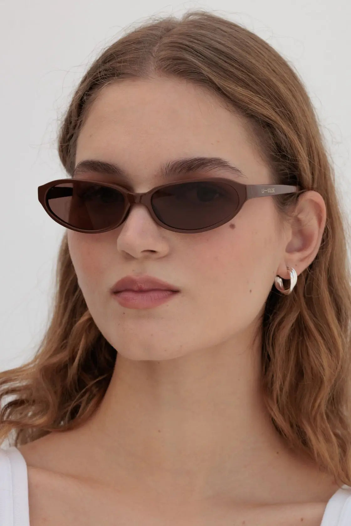 Model wears cinnamon oval-frame sunglasses styled with soft waves, perfect for warm-weather simplicity or layered neutrals.