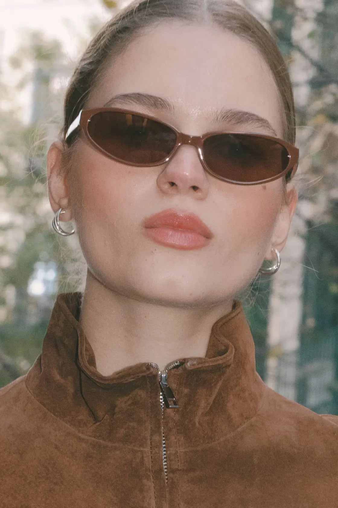Model in cinnamon-toned sunglasses and a tan suede jacket, set against a soft natural backdrop—perfect for autumn layering or cool-weather escapes.