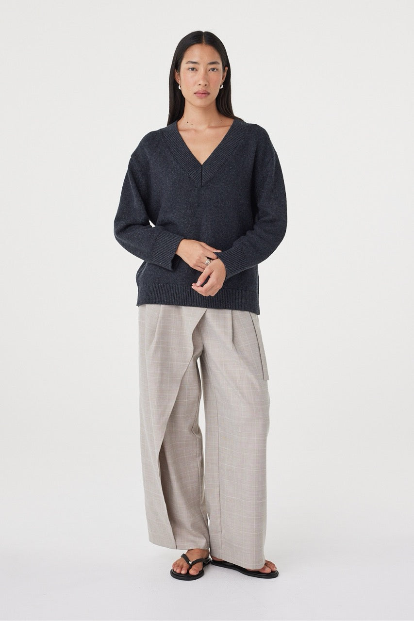 Full-length view of the Zenya Sweater in Dark Grey Marle with dropped shoulders, ribbed trims and an easy oversized fit, paired with light grey trousers and sandals.