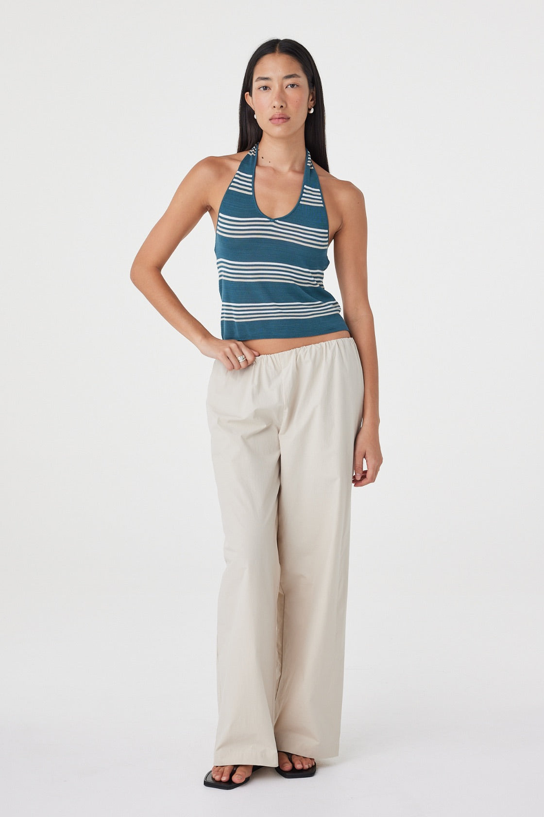 Full-length front view of the Nuri Top in Teal Stripe styled with cream pants, showing the halterneck shape and close fit.