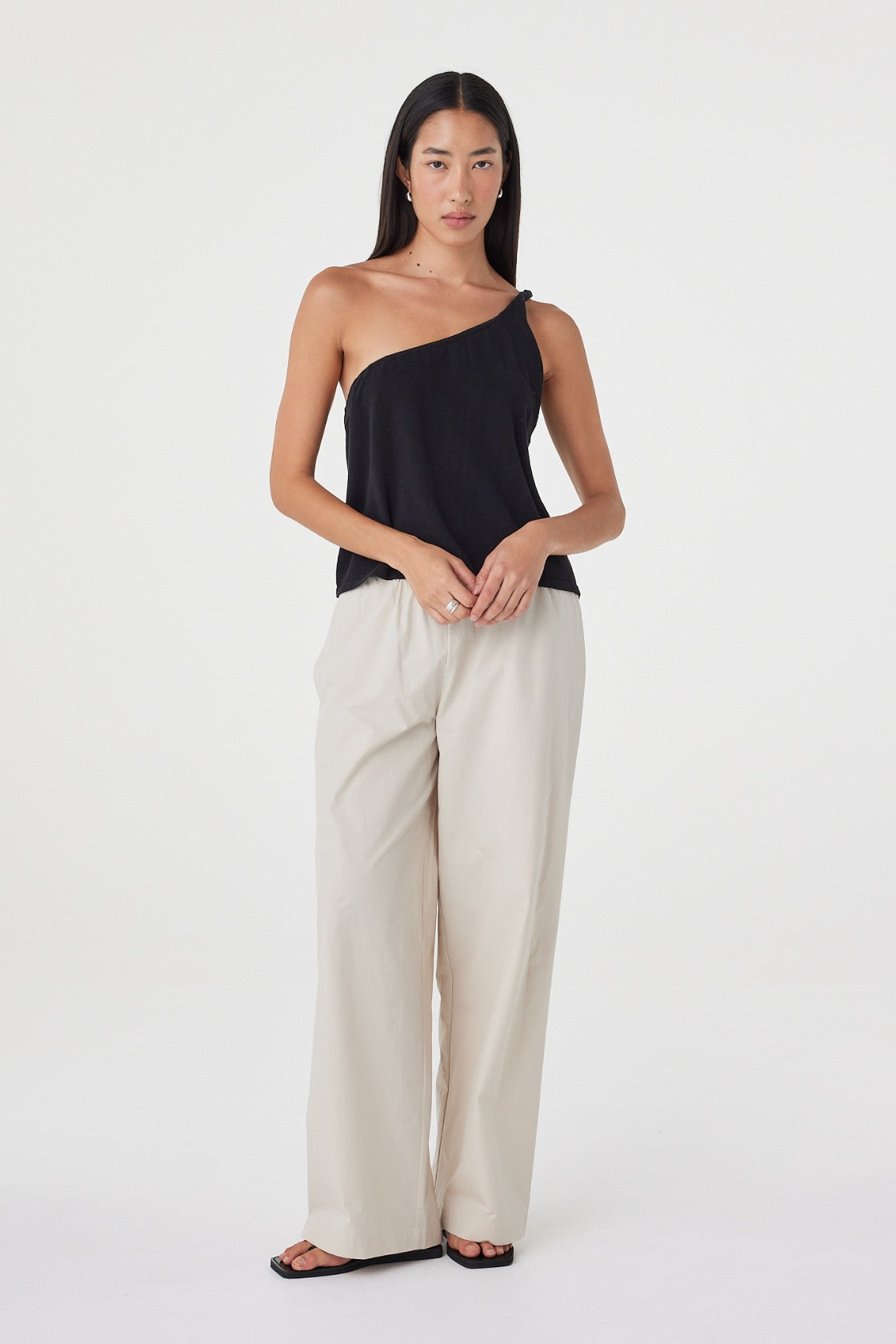 Full-length front view of the Harri Asym Top in Black showcasing the one-shoulder design and softly draped fit, paired with taupe wide-leg pants