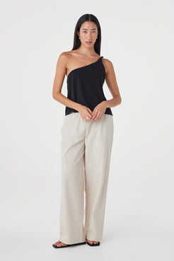 Full-length front view of the Harri Asym Top in Black showcasing the one-shoulder design and softly draped fit, paired with taupe wide-leg pants