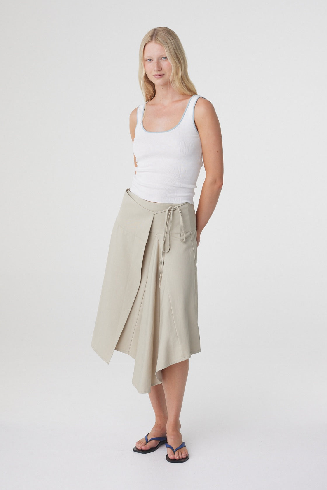 Anya Skirt in Taupe, relaxed three quarter pose styled with a white tank and flat sandals, showing the movement and asymmetric shape of the skirt.