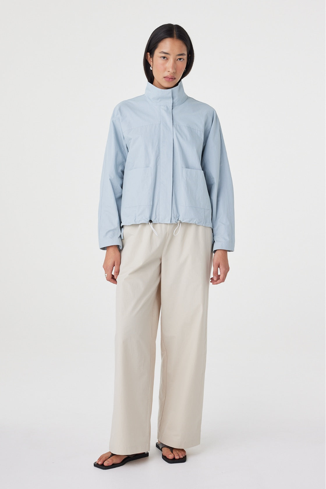 Angled front view of the Addie Jacket in Pale Blue featuring the funnel neckline, oversized patch pockets and adjustable toggle hem, styled with wide-leg trousers.