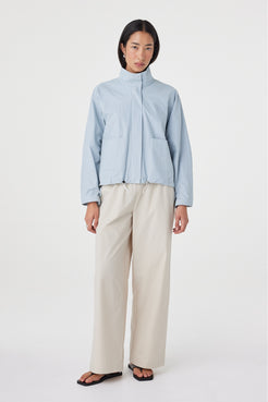 Angled front view of the Addie Jacket in Pale Blue featuring the funnel neckline, oversized patch pockets and adjustable toggle hem, styled with wide-leg trousers.