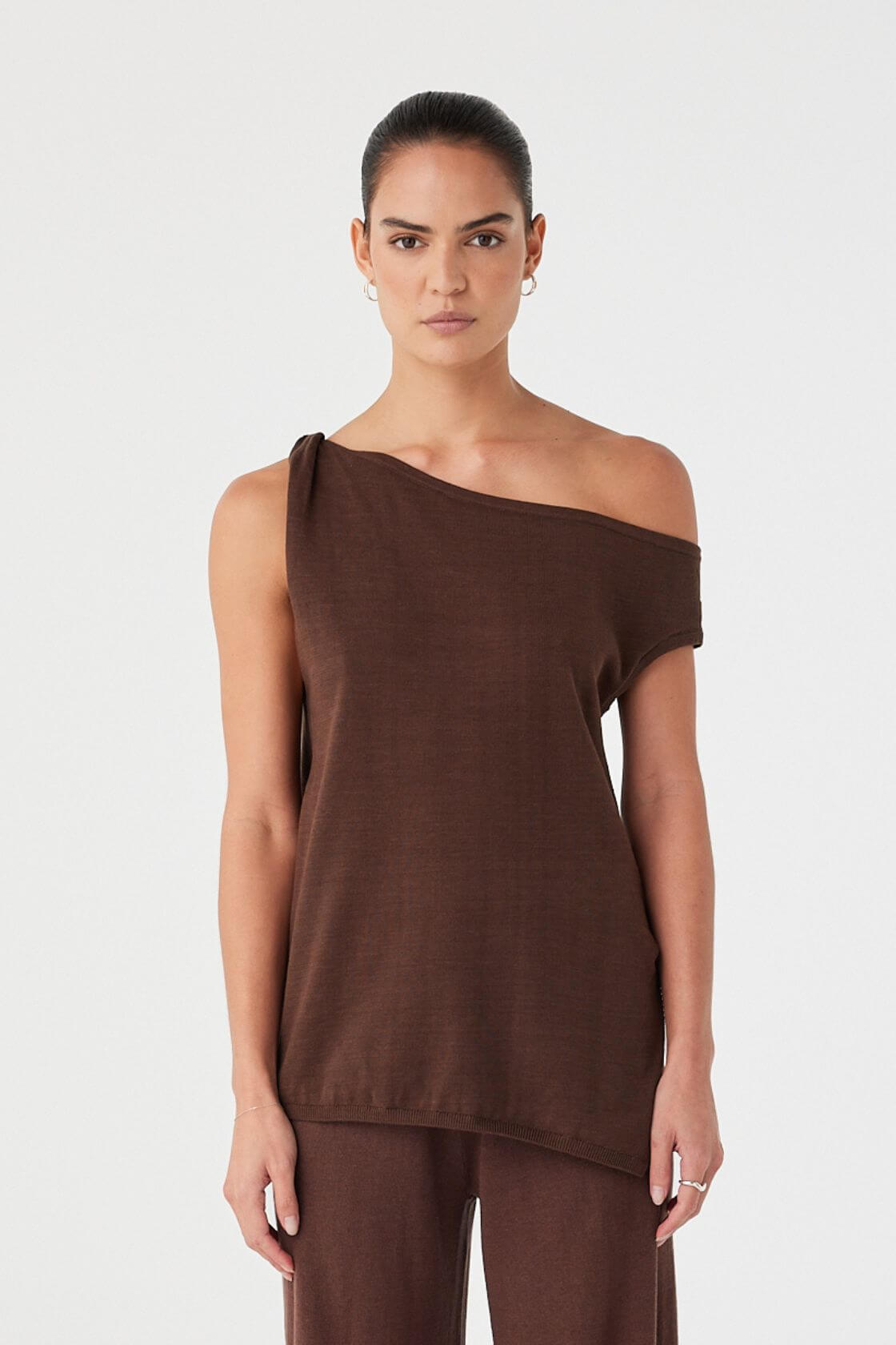 Model wearing brown cotton knit asymmetric top with off-shoulder neckline, side twist, and relaxed longline fit.