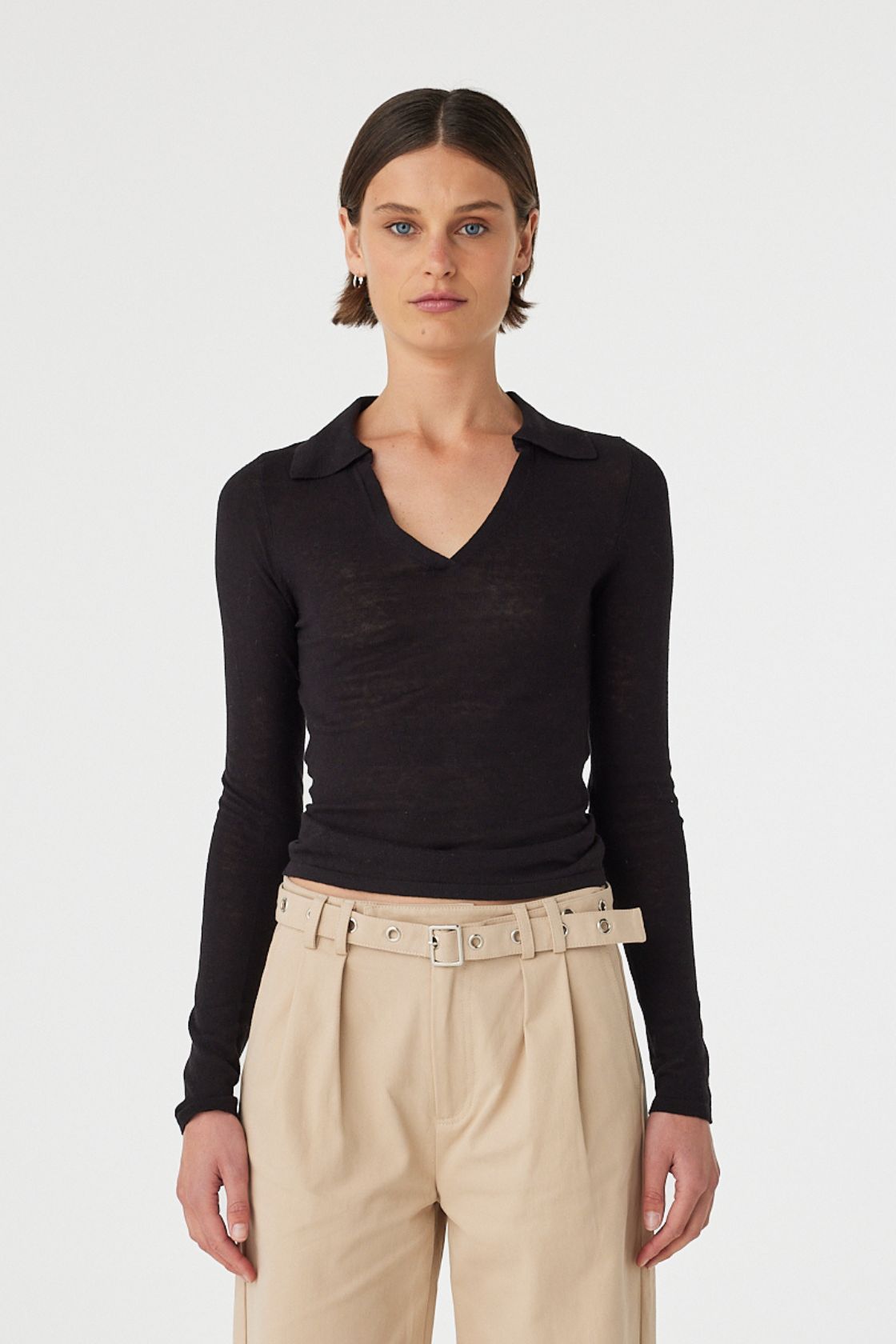 Close up of Mae longsleeve top in black worn front on, showcasing the slim fit silhouette, semi sheer knit texture and V neckline with polo collar, styled with beige wide-leg pants 