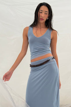 A woman modeling a light blue two-piece set consisting of a sleeveless V-neck crop top and a matching high-waisted maxi skirt, styled with a dark beaded belt featuring a silver ring, against a draped white fabric backdrop.