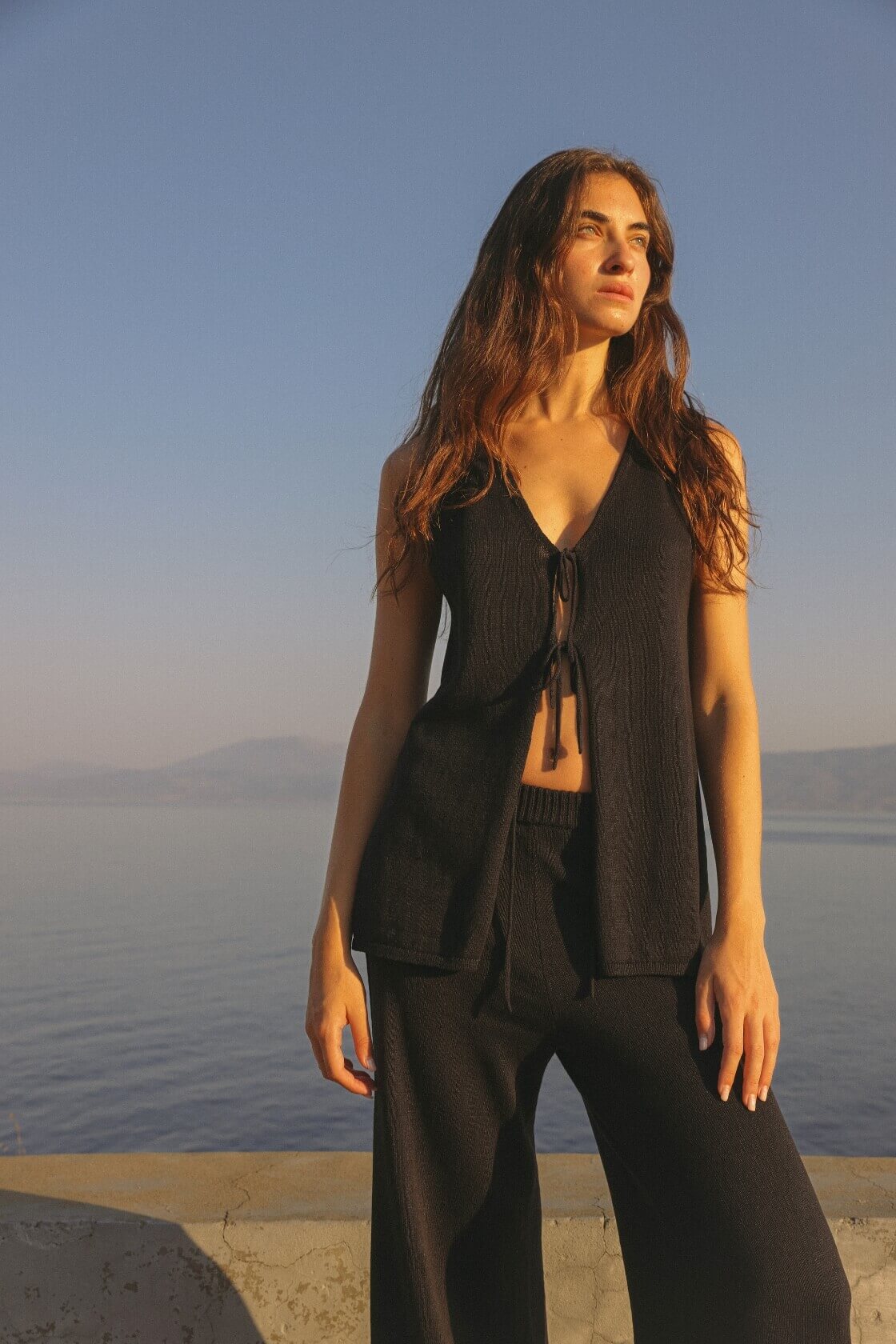 Model wears black reversible longline top with front tie closure and deep V neckline, styled with matching wide-leg pants—soft knit layers at golden hour.