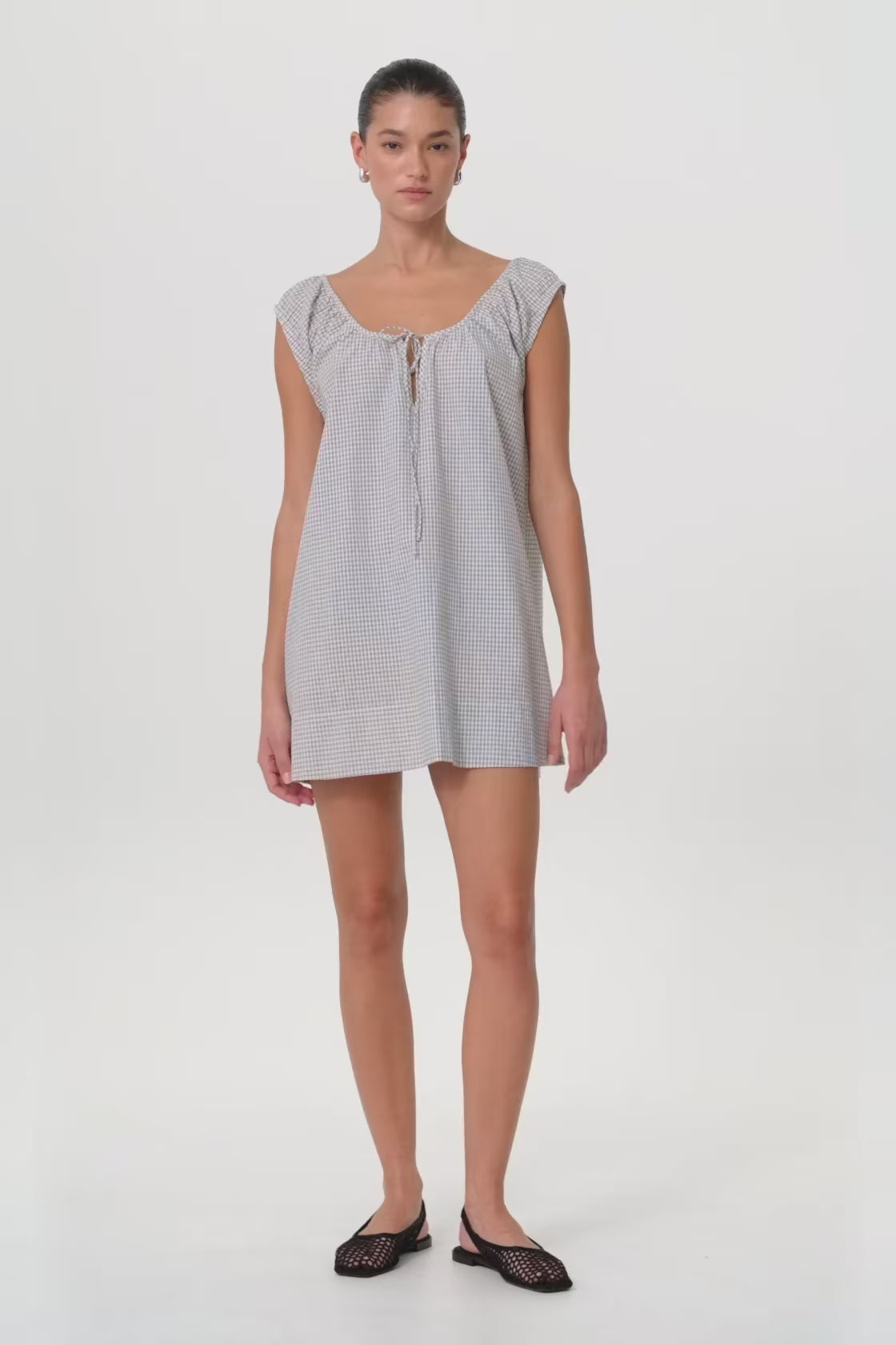 Model in motion wearing a flared cotton mini dress with adjustable neckline and soft check texture, designed for ease through changing seasons.